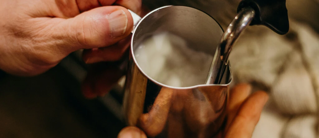 Barista using a steam wand to froth milk in a metal jug, carefully creating a smooth and creamy texture necessary for specialty coffee drinks.