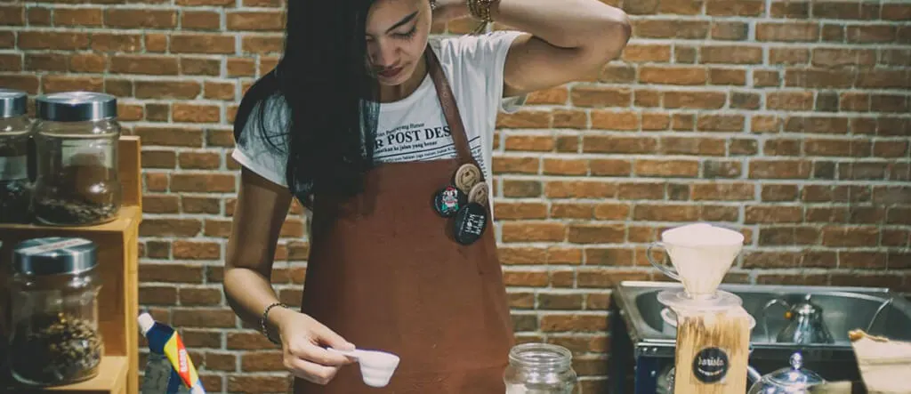 A female barista holding a small milk server, preparing to froth milk for a latte or cappuccino.