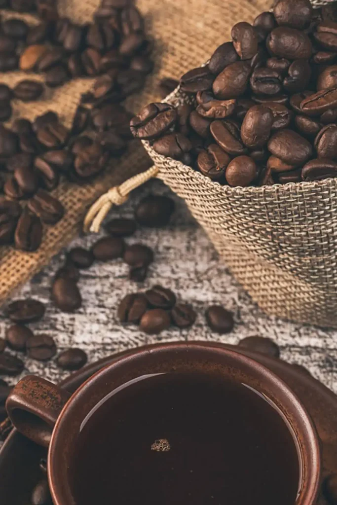 A rustic hessian bag filled with coffee beans next to a steaming cup of black coffee, highlighting freshness and bold flavor.