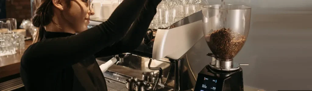 ☕ Conical vs Flat Burr Grinders: What’s the Difference? 183 Woman pouring coffee beans into a grinder in a modern kitchen setting.