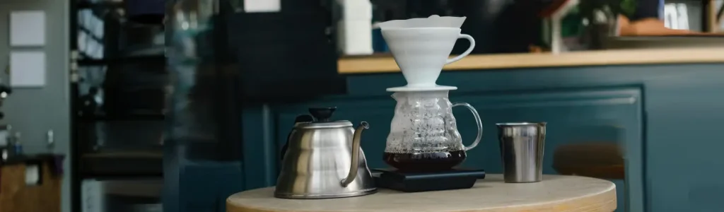 How to Drip Coffee (Step-by-Step) 48 Hario V60 dripper brewing coffee into a glass carafe, with a kettle and coffee cup beside it.
