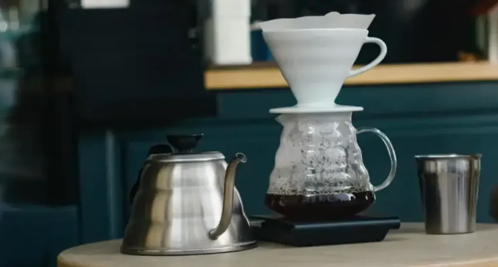 How to Brew Hario V60 Coffee Like a Pro ☕ 65 Hario V60 dripper showing how to brew Hario V60 Coffee into a glass carafe, with a kettle and coffee cup beside it.