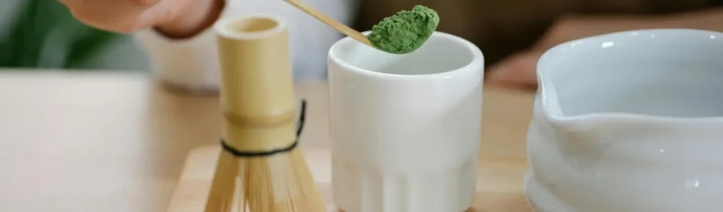 How to Make the Perfect Matcha Latte at Home 82 Bamboo whisk and matcha powder being poured into a coffee cup to make a frothy matcha latte at home.
