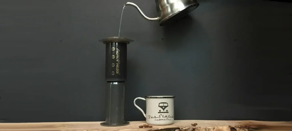 Portable Coffee Makers for Travel (Ultimate Guide) 96 Person pouring hot water from a kettle into an AeroPress coffee maker with a cup beside it