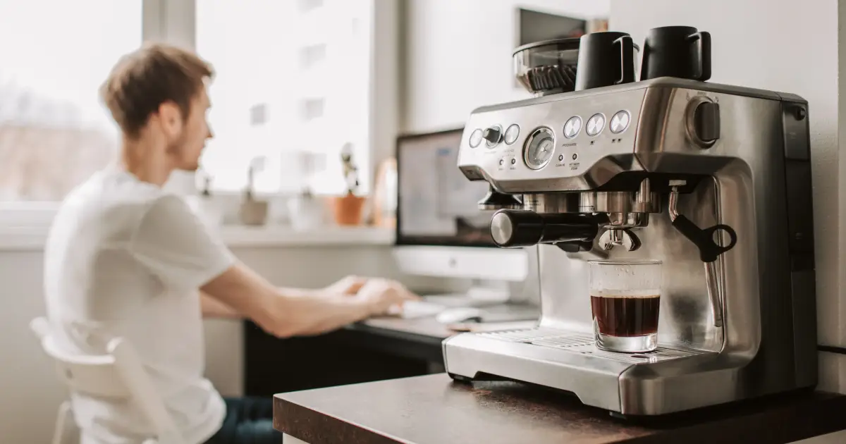 How to Choose a Coffee Machine for Your Home 2 Young man researching how to choose a coffee machine for your home while sitting beside an espresso machine in a modern kitchen.